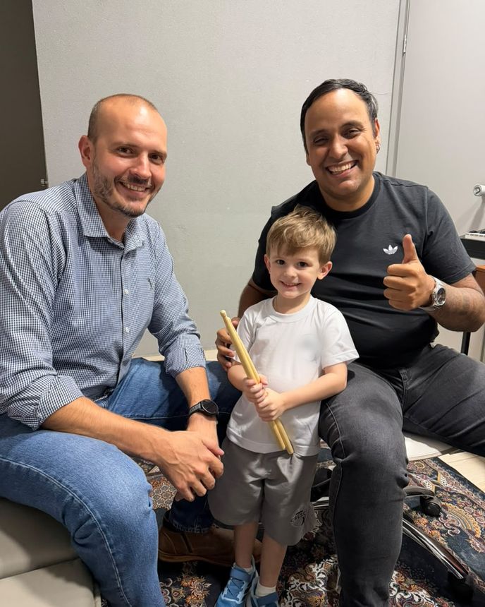 Dois adultos e um menino com baquetas na mão, sorrindo na sala da escola