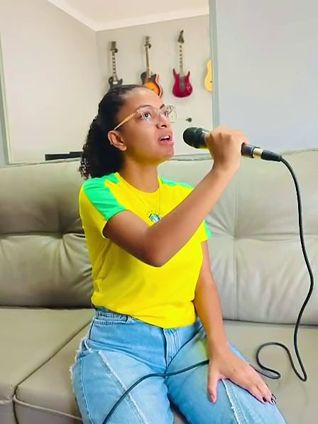 Aluna de óculos e camiseta amarela cantando ao microfone, com guitarras na parede ao fundo