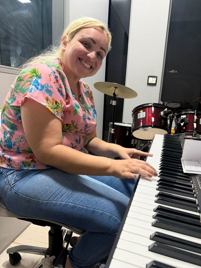 Aluna adulta sorrindo ao teclado, com a bateria ao fundo